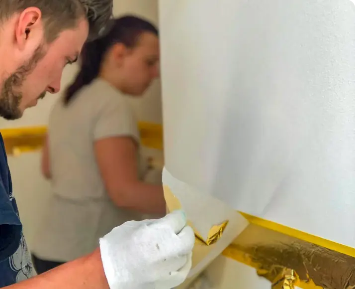 Handwerker trägt mit Handschuhen eine goldene Verzierung auf eine weiße Wand auf, während eine weitere Person im Hintergrund arbeitet.“